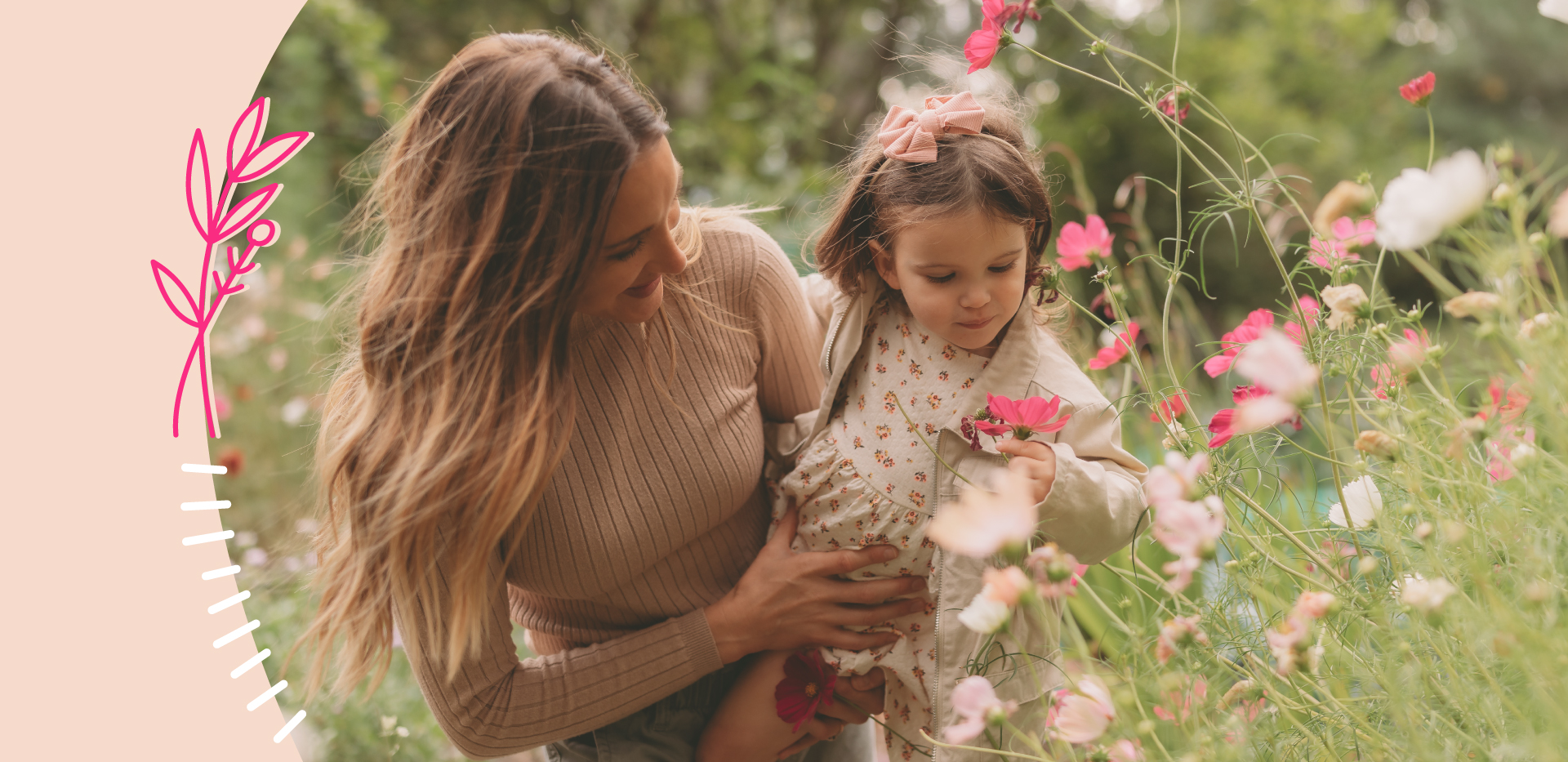 The image shows a woman with brown hair holding a little girl with brown hair and a dress. They are standing in a field of tall grass and flowers. The woman is smiling, and they are both looking down at the pink and white flowers in front of them, towards the bottom right of the image.